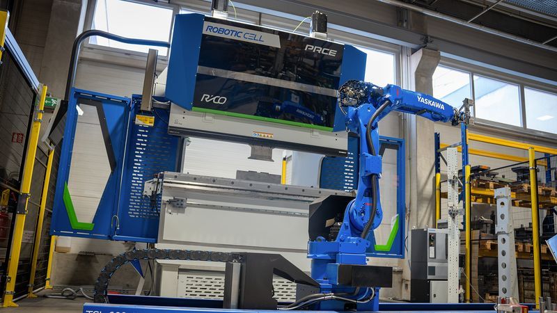 A blue industrial robot arm is positioned next to a large blue and green metal fabrication machine.