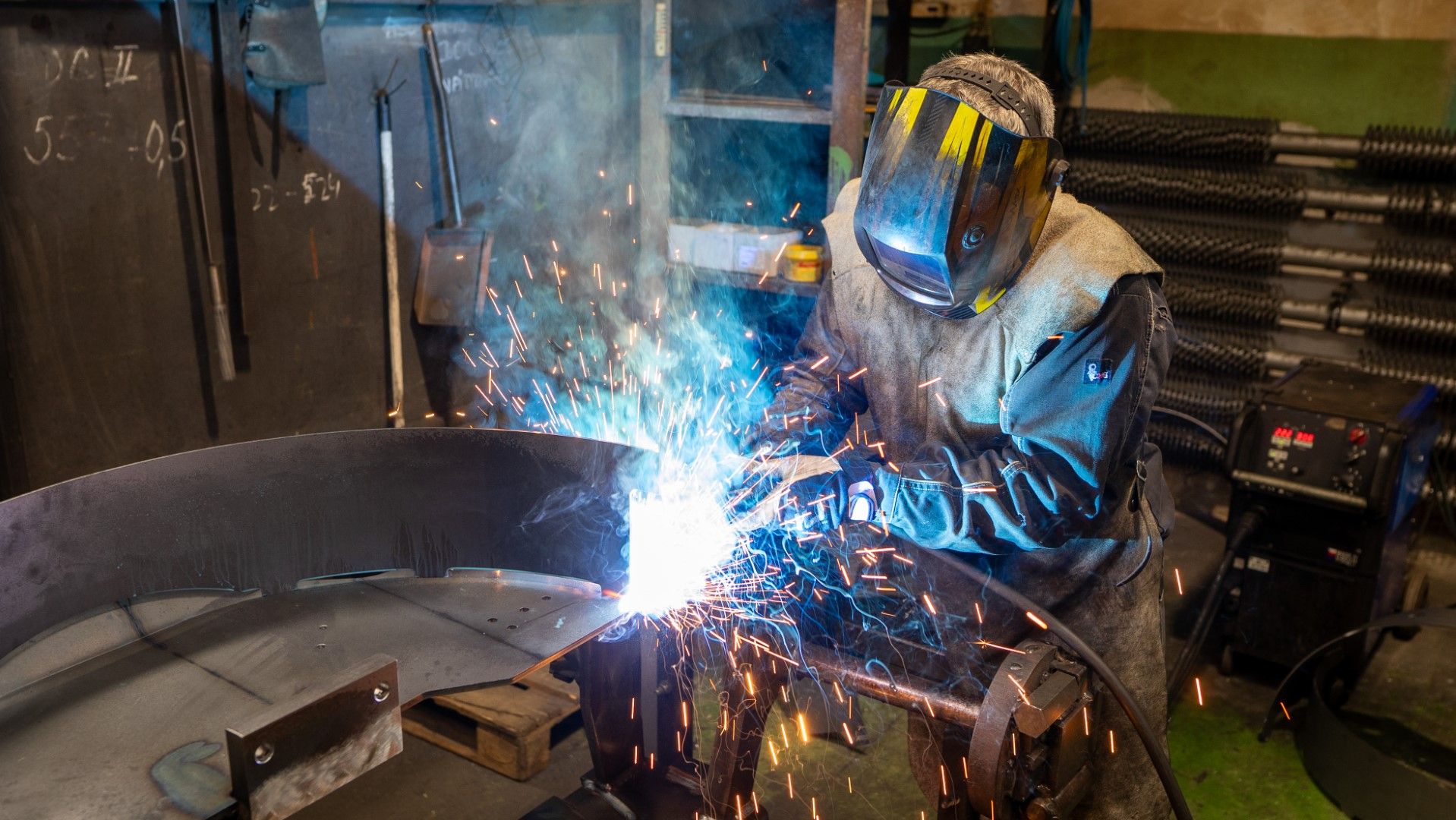 A welder wearing protective gear works on a large metal object, creating a bright spark shower.