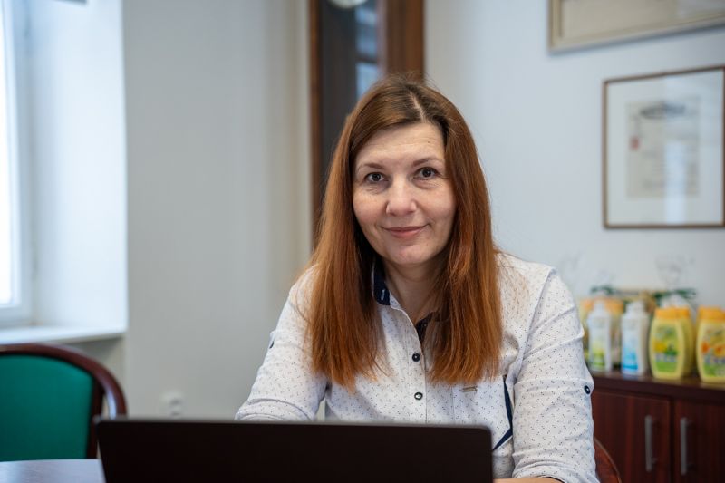 Red-haired woman in white shirt smiles, sitting at an office desk with a laptop.