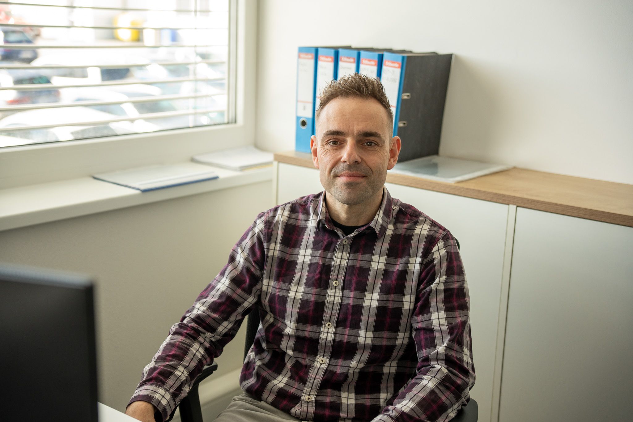 A man with short hair and a plaid shirt sits at a desk, smiling.