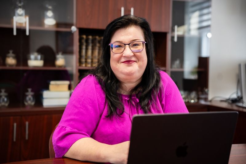 Smiling woman with dark hair and glasses in magenta top at a desk.