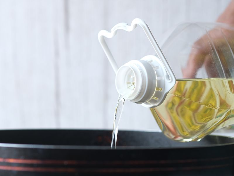 A person pours golden cooking oil from a clear plastic bottle into a dark frying pan.