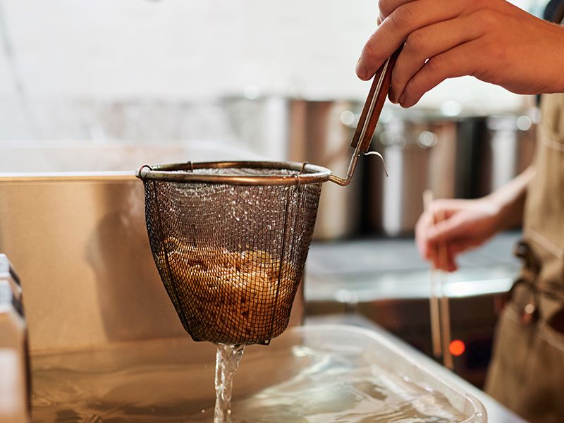 A hand lifts a metal strainer full of steaming noodles from boiling water in a kitchen.
