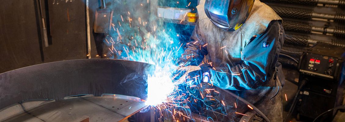 A welder in protective gear creates bright sparks while working on metal in a dimly lit workshop.